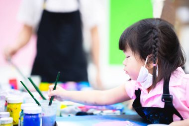 Kız sınıfta sanat yapıyor. Çocuk fırçayı bir bardak suda yıkıyor. Çocuklar çenelerine bez bir maske takarlar. Çocuk suda resim yapmayı öğreniyor. Bebek 3-4 yaşında..