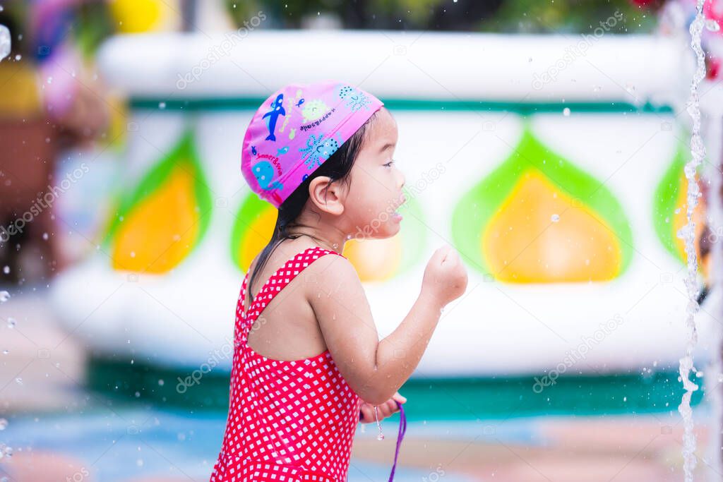 Selective focus. Portrait little girl was agape the fountain pouring out from the ground in front of her. Child make funny faces. Active kid 3-4 years old in a red bathing suit and a pink swim cap.