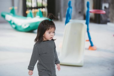 Little girl in a moody mood. Children peck their fingernails. Background is a playground, simulating a small playground in the area of the house. Child aged 3-4 years old.