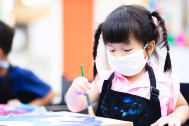 Öğrenci kız, tuvalde fırça ve suluboya ile resim yapmaya odaklanmıştı. Boyamayı öğrenirken beyaz bez bir maske takan küçük Asyalı çocuk. 3 yaşındaki çocuklar.