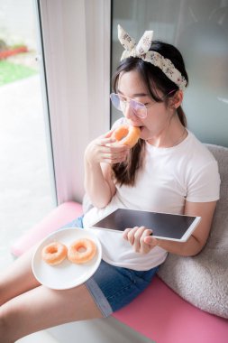 Beyaz tişört ve gözlük takan Asyalı genç bir kadın evden tablet dokunmatik ekranda çalışıyor. Ve rahatça kanepede donut yiyebilirsin..
