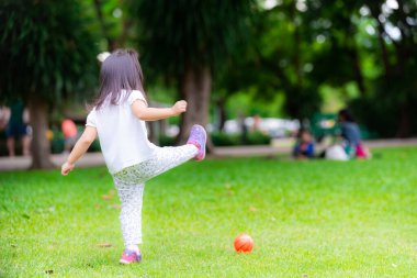 Çocuğun arkası, yeşil çimlerde küçük turuncu topları tekmeleme oyunu oynuyor. Beyaz gömlek giyen ve spor yapan Asyalı bir kız. 3 yaşındaki çocuk egzersiz yapıyor..