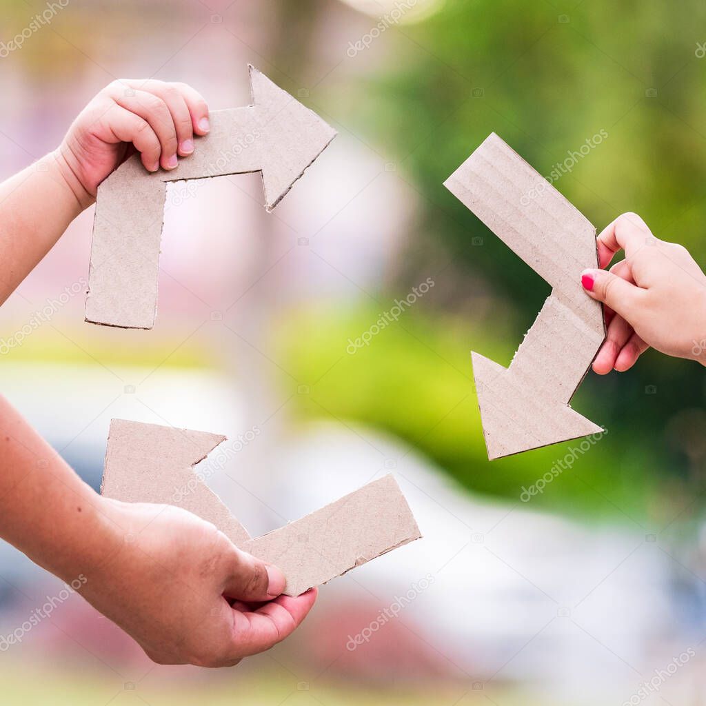 The hands of people holding arrow symbols cut from old light brown paper boxes are shown as reused symbols against nature backgrounds. Ecological concepts.