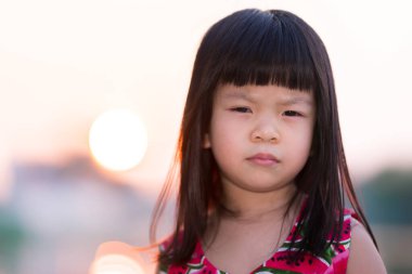 Asyalı kızın yüzüyle yakın çekim duygusal ifadesi. Çocuk tuhaf bir yüz göstererek kaşlarını çattı. Parkta günbatımı arka planı. Doğal Fuar. Rim Light. 4 yaşında çocuklar..