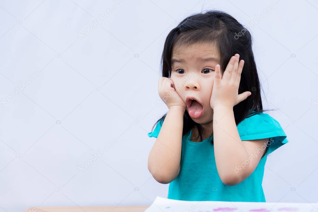 Child appears shocked or bored. Children pushes her cheek with both hands. Kids sticks out his tongue, makes eyes big. Drawing book was placed on wooden table in front of her. Girl is 3-4 years.