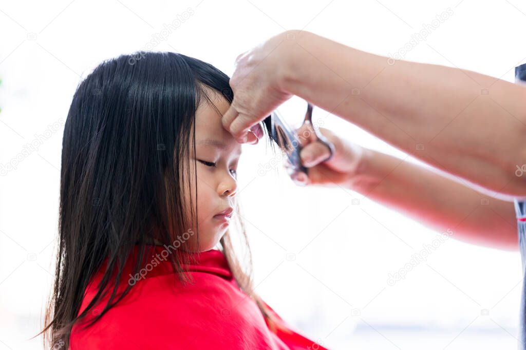 Asian little girl sitting haircut at the beauty salon. Cute child cut the bangs. Happy kid covered a red cloth. The barber's hand is skillfully cutting the hair. Children is 4 years old.