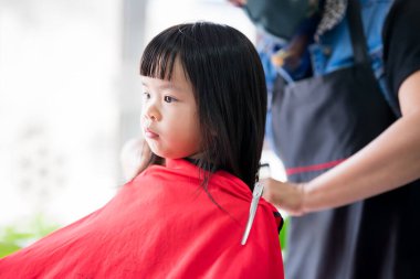 Asyalı kız, kuaförde saçını kestiriyor. Küçük çocuklar için saç kesimi. Çocuk, saçın kıyafetlere girmesini önlemek için cesedi kırmızı peçeyle örtüyor. Çocuklar 4 yaşında..