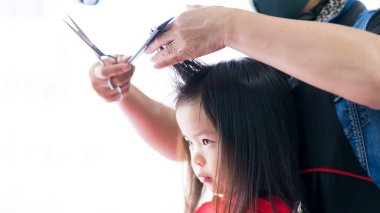 Seçici odaklanma. Kuaför, çocuk kıllarını kesmek için onun uzman ellerini kullanıyordu. Ön tarafı kesmek için küçük kızın saçını tutuyordu. Kuaförün ellerinde, ustalıkla tutulmuş bir makas ve bir tarak.