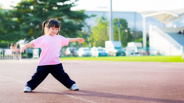 Sevimli Asyalı kızlar egzersiz yapıyor. Mutlu çocuk tatlı tatlı gülümser. Hoş çocuklar stadyumda spor yapıyor. Çocuklar kollarını açar. Yaz ya da bahar akşamları. Pembe tişört giyen bebek 4 yaşında..