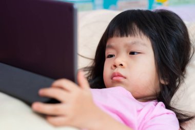 Kızı yatağında yatıp tabletten çizgi film izliyordu. İnternetle videodan öğrenen çok tatlı bir kız. Asyalı çocuk öğrenmek ve eğlenmek için dokunmatik ped kullanır. Pembe gömlek giyen 4 yaşında bir çocuk..
