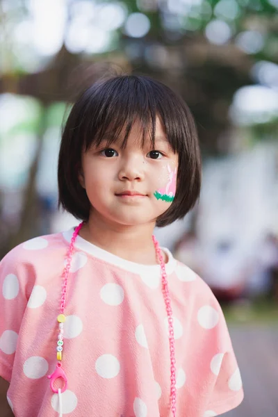 Cute Asian girl with sweet smile looking at camera. Child wear pink shirt. Kid\'s face was painted with watercolors. Sand flakes trace the children\'s face. Black-haired lady aged 5-6 years. Easter day.