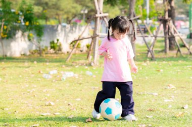 Yeşil çimlerde mavi beyaz futbol topu tekmeleyen aktif, sevimli Asyalı kız. Sıcak ve güneşli bir günde egzersiz. Yazın ya da ilkbaharda. Mutlu 4 yaşındaki çocuk egzersiz kıyafeti giyiyor..