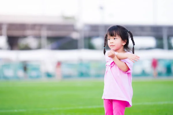 可愛いアジア系の女の子が草の中で運動している 幸せな子供は両腕を使用しており 左右の体をねじっています 子供たちはスポーツを楽しむ ピンクのシャツを着た４歳の子供 — ストック写真