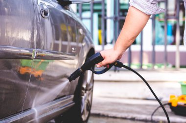 Adamın eli siyah, yüksek basınçlı bir hortumu tutuyor, arabayı temizliyor. Su damlaları saçıldı..