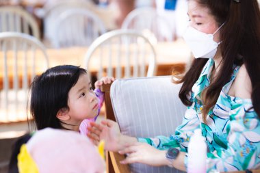 Asyalı anne denize girmeden önce kızına güneş kremi sürüyor. Cildini ultraviyole ışınlardan korumak için. Annem beyaz bir maske takıyor. Dinlenme günü. Tatil.
