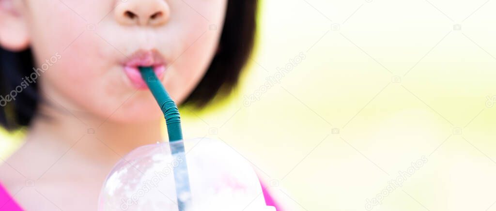 Cropped imaged. Asian child girl sucking water from a plastic glass with a green straw. Cinematic ratio.