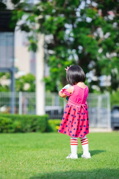 垂直成像 回顾一下亚洲女孩站在绿色法律上的景象 孩子手里拿着球 穿着红白相间的长袜的孩子 夏天的儿童运动 5岁的粉红婚纱婴儿 — 图库照片