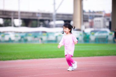 Kızıl stadyumda koşan Asyalı aktif bir kız. Yazın ya da ilkbaharda. Akşamları çocuk egzersizi. Pembe tişörtlü, bacaklı, beyaz ayakkabılı bir çocuk. 4-5 yaşlarında mutlu çocuklar..