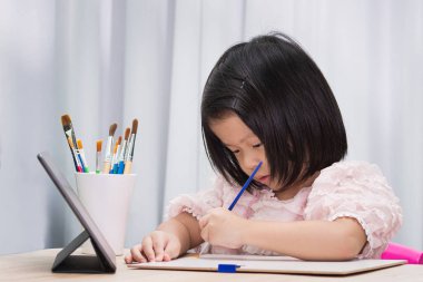 Çevrimiçi okul konsepti. Kağıt üzerinde suluboya boyalarla resim yapan Asyalı tatlı kız. Çocuk dokunmatik yere bakıyor. Çocuk ahşap masada resim öğreniyor. 4-5 yaşlarında güzel bir bebek..