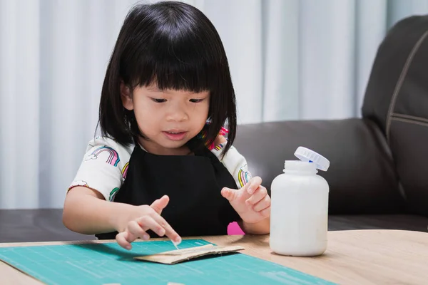 Sweet child girl applying pieces from cardboard boxes using glue while doing arts and crafts at lesson classroom in school. Crafts for kids. Project of kid\'s creativity. Handicrafts work.