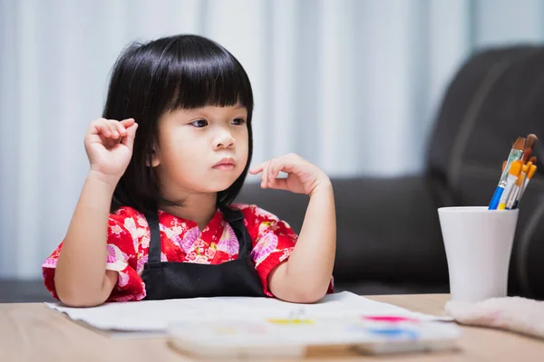 Asian girl sits and thinks, imagining the topic of art that will be done today. Child are sitting gazing out and making plans for tasks to do. Kid wearing black apron as her to work on watercolor art.