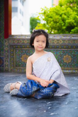 Geleneksel Tayland giysisi Sabai ve gümüş renkli parlak ayakkabı ve broş takan Asyalı kız çocuğunun güzel geleneksel portresi. Çocuk kibarca taştan zeminde oturuyor..