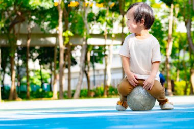 Mutlu bir çocuk parkta güneşli bir günün keyfini çıkarır, geniş bir gülümsemeyle yıpranmış bir futbol topunun üzerinde dinlenir. Canlı ortam ve eski top basit, aktif açık hava eğlencesini ve spor tutkusunun başlangıcını simgeliyor.