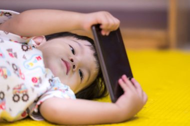 Sarı bir paspasın üzerinde oturan genç bir Asyalı çocuk, dikkatle akıllı telefon ekranına odaklanmış. Erken dijital pozlama kavramı, yeni yürümeye başlayanlar için ekran zamanı ve evdeki modern çocukluk yaşam tarzı.