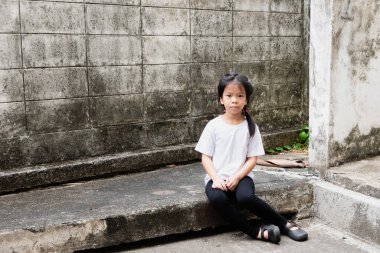 Yalnız bir Asyalı genç kız beton bir çıkıntıda eski, yıpranmış tuğla bir duvara karşı oturuyor. Kentsel ortamlarda sosyal eşitsizlik, çocukluk yoksulluğu, ihmal edilen gençlik ve çocuk refahı kavramı.