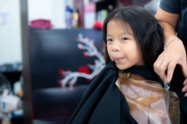 Profesyonel bir kuaförde saç tıraşı olan yakışıklı bir Asyalı kız. Siyah bir pelerinle kaplı bir şekilde berber koltuğunda oturan bir çocuğun portresi..