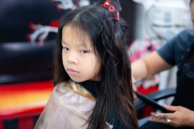 Saçını kestirirken yan bakışlarından sıkılmış ve rahatsız olmuş genç bir Asyalı kızın yakın çekimi. Çocukluk sabırsızlık anlayışı, uzun güzellik salonu seansları ve gerçek çocukların yüz ifadeleri..