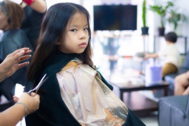 Genç bir Asyalı kızın modern bir kuaförde saçını kestirip doğrudan kameraya bakarken orta boy fotoğrafı. Kopya alanı olan kentsel yaşam tarzında çocuklar için profesyonel saç bakım hizmeti.