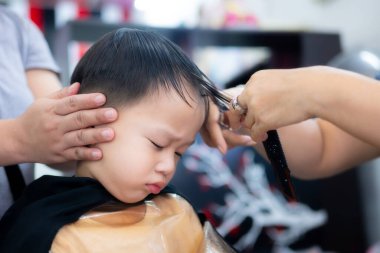 Bir kuaförde saçını kestirirken gözlerini kapatan uykulu bir Asyalı çocuğun yakın çekimi. Stilistler ellerini nazikçe başını destekliyor. Bakım sırasında çocukluk yorgunluğu ve sabrı kavramı.