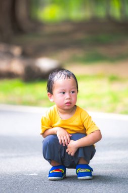 Sarı tişörtlü, parkta çömelmiş meraklı bir gencin samimi dikey portresi. Yumuşak açık hava aydınlatması ve yemyeşil bokeh arka plan masum bir çocukluk keşfini vurguluyor..