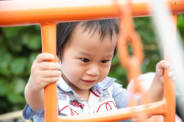 Çocuk parkında turuncu metal çerçeveye tırmanan kararlı bir çocuğun yakın plan portresi. Bir anlık odaklanma, fiziksel koordinasyon ve erken çocukluk keşiflerinin masumiyetini yakalamak..