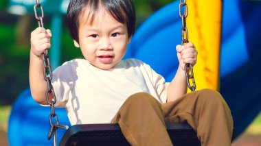 Siyah sallanan koltukta oturan genç çocuk, oynarken metal zincirleri sıkıca tutmak için iki elini kullanıyor. Açık hava çocuk parkında renkli bir geçmişi olan samimi bir çocukluk aktivitesi..