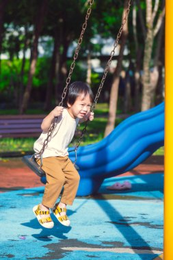 Beyaz tişörtlü, kahverengi pantolonlu, açık hava çocuk parkında salıncaktan hoşlanan neşeli küçük bir çocuğun tam boy portresi. Çocuk sallanırken metal zincirleri kavrıyor, mavi kaydırak ve yeşil ağaçlar arka planda