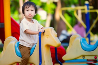 Açık hava çocuk parkında sarı yay binicisi atının üstünde oturan krem rengi tişörtlü çocuk. Çocuk, arka planda bulanık oyun alanı ekipmanları ve yeşil ağaçlarla, tuhaf bir ifade..