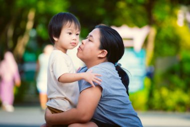 Annenin portresi çömelmiş, oğlunu kucaklıyor ve ona yanağından sevgi dolu bir öpücük vermek için eğiliyor. Parktaki hassas an, anne ve çocuğun derin duygusal bağlarının özünü yakalar.