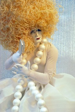 Beyaz elbiseli, beyaz eldivenli oyuncak bebek sarı saçlarını tarıyor.