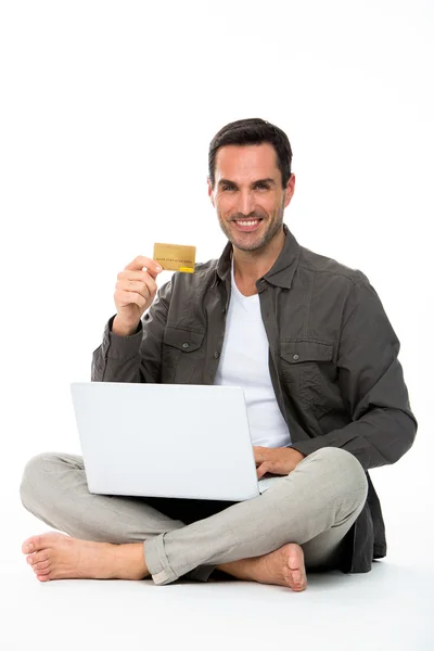 Hombre sentado en el suelo, sonriendo a la cámara, mostrando la tarjeta de crédito y la compra en línea — Foto de Stock