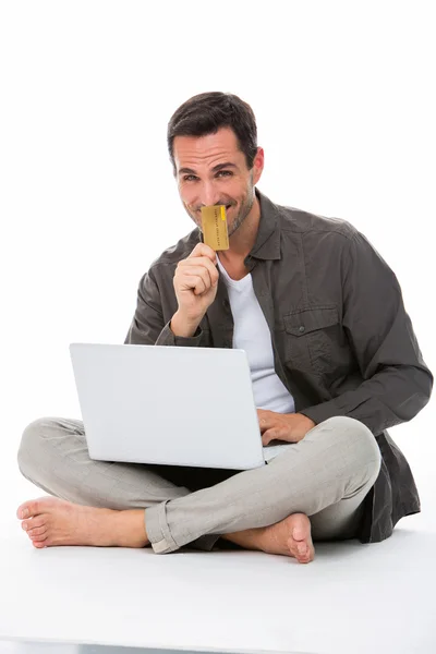 Hombre sentado en el suelo, sonriendo a la cámara, sosteniendo la tarjeta de crédito y comprando en línea — Foto de Stock