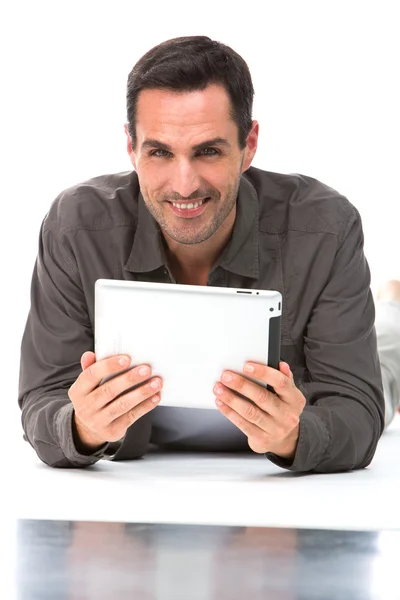 Hombre tendido en el suelo, sonriendo a la cámara y sosteniendo su tableta digital con ambas manos — Foto de Stock
