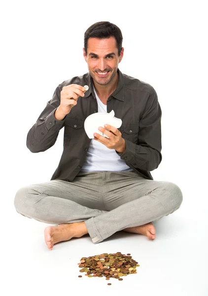 男人坐在地板上，微笑着摄像机，拿着一枚硬币放在储钱 — 图库照片