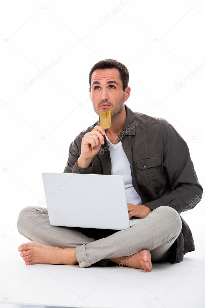 Thoughtful man sitted on the floor holding credit card and buying online
