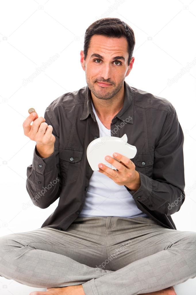 Man sitted on the floor, looking at camera, putting a coin in a piggybank