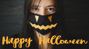 Siyah maskeli genç bayana Cadılar Bayramı mesajınız kutlu olsun. Koyu renk ahşap arka planda Jack O Lantern gülüşü. 2020 Cadılar Bayramı, Coronavirus sırasında güvende olun. El yazısı, mevsimlik tebrik kartı.