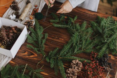 Elleri makas ve yeşil dallar tutan kadın eller, ahşap masanın arka planında Noel çelengi yapıyor. El yapımı şenlik dekoru. Kırsal Noel çelengi yapmak, mevsimsel kış gelişimi