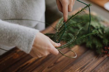 Yeşil köknar dallarını tutan kadınların ellerini kapat ve ahşap masanın arka planında rustik Noel çelengi yap. El yapımı şenlik dekorasyonu, tatil gelişimi. Mevsimlik kış atölyesi