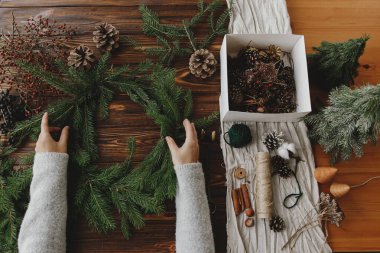 Kırsal Noel çelengi düzleştiriyor. Çam kozalakları, böğürtlenler ve doğal şenlik süslemeleri olan ahşap masa üzerinde yeşil köknar dalları tutan eller. Mevsimlik kış atölyesi, tatil gelişimi